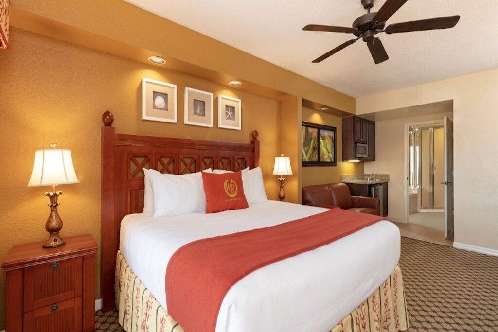 Habitación de hotel con una cama grande, dos lámparas de noche, un ventilador de techo, un escritorio pequeño y una puerta que da a un baño.