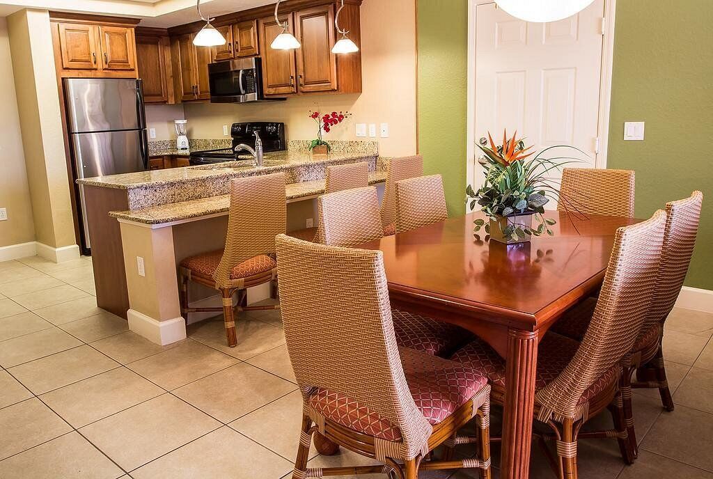 Cocina con armarios de madera, refrigerador de acero inoxidable y encimera de granito. Hay una mesa de comedor con sillas de mimbre y una planta en maceta.