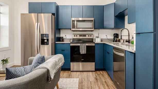 Une cuisine moderne avec des meubles bleus, des appareils en acier inoxydable et un canapé gris dans le salon attenant.