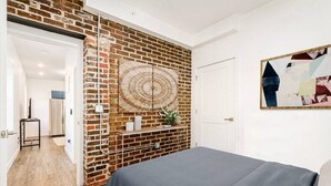 Une chambre avec un mur en briques, un lit recouvert d’une couverture grise, une étagère fixée au mur ornée d’objets décoratifs et une porte donnant sur une autre pièce.