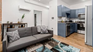 Una sala de estar moderna con un sofá gris, un tapete estampado y una cocina con armarios azules y electrodomésticos de acero inoxidable.