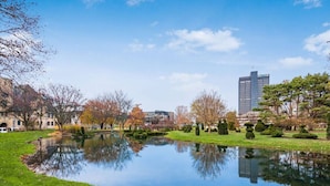 A serene park with a pond, manicured lawns, and a modern building in the background. - Central Loft with Pool Table! Pet-friendly Haven! (Columbus)