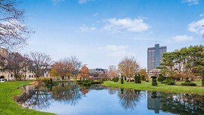 A serene park with a pond, manicured lawns, and a modern building in the background. - Modern Apartment with Pool Table! Pet-friendly! (Columbus)