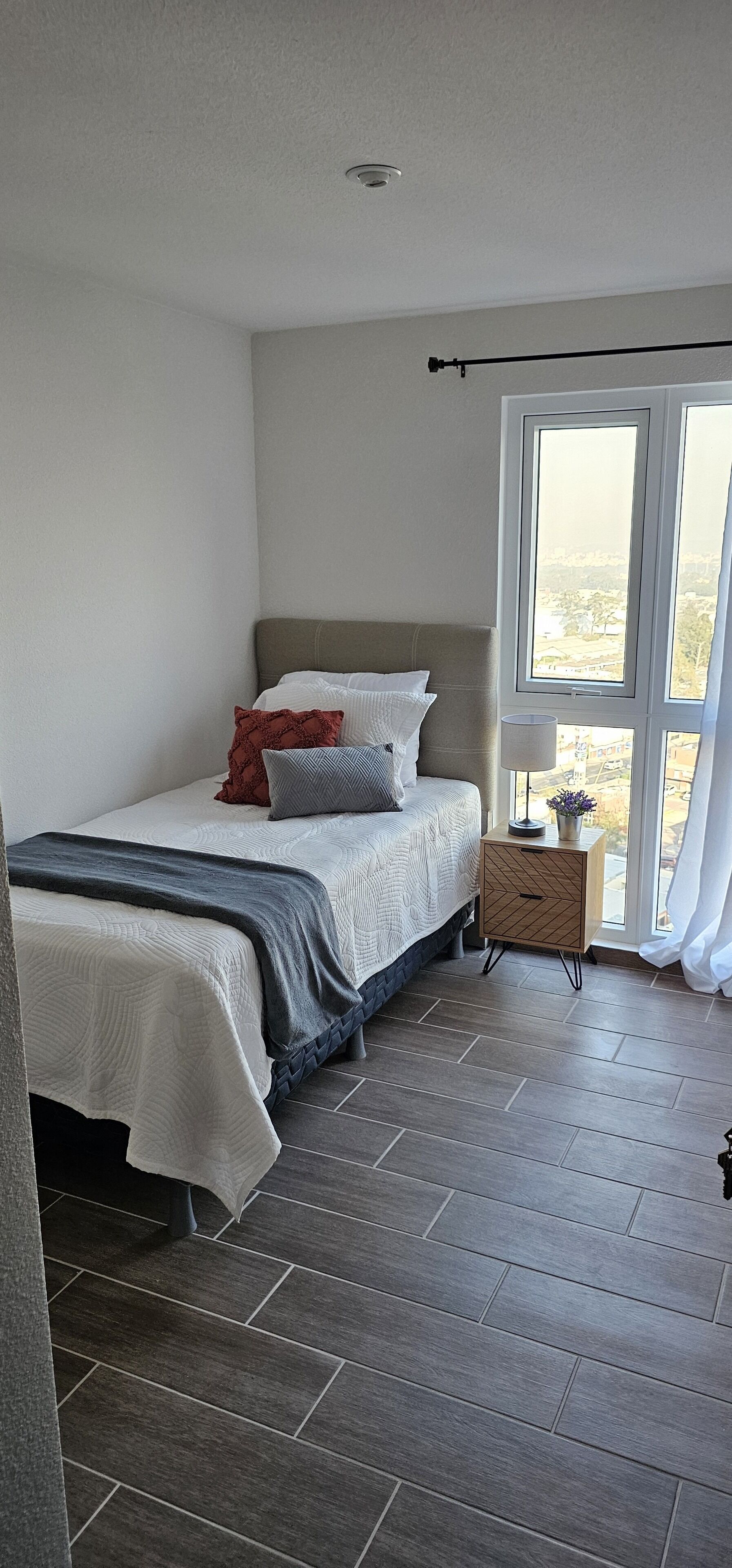 Un dormitorio con cama, mesita de noche con lámpara y ventana con cortinas.