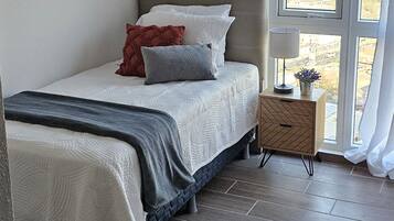 Un dormitorio con cama, mesita de noche con lámpara y ventana con cortinas.