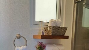 Un baño con inodoro, ducha y toallero con una toalla blanca. Hay estantes de madera sobre el inodoro que sostienen una cesta, una planta y papel higiénico.