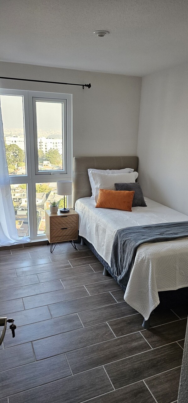 Une chambre à coucher avec un lit, une table de chevet, une lampe et une vue sur l’extérieur grâce à une grande fenêtre.