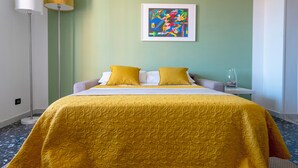 A hotel room with a bed, a floor lamp, a framed abstract painting, and a small table with a vase. - Per una vacanza meravigliosa. (La Spezia)
