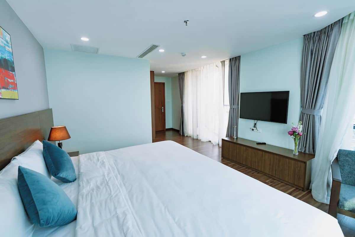 Una habitació d'hotel amb un llit gran, un televisor penjat a la paret, un panell de fusta al capçal del llit, una làmpada de nit i una finestra amb cortines.