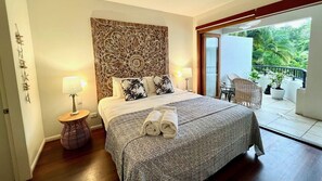 Ein Hotelzimmer mit einem großen Bett, zwei Nachttischlampen, einem dekorativen Kopfteil, einem Balkon mit Sitzgelegenheiten und Blick auf Grünflächen.