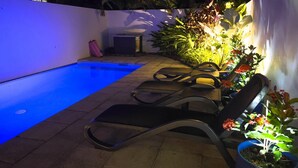 Ein Swimmingpool mit blauer Beleuchtung, Liegestühlen und Topfpflanzen am Rand.