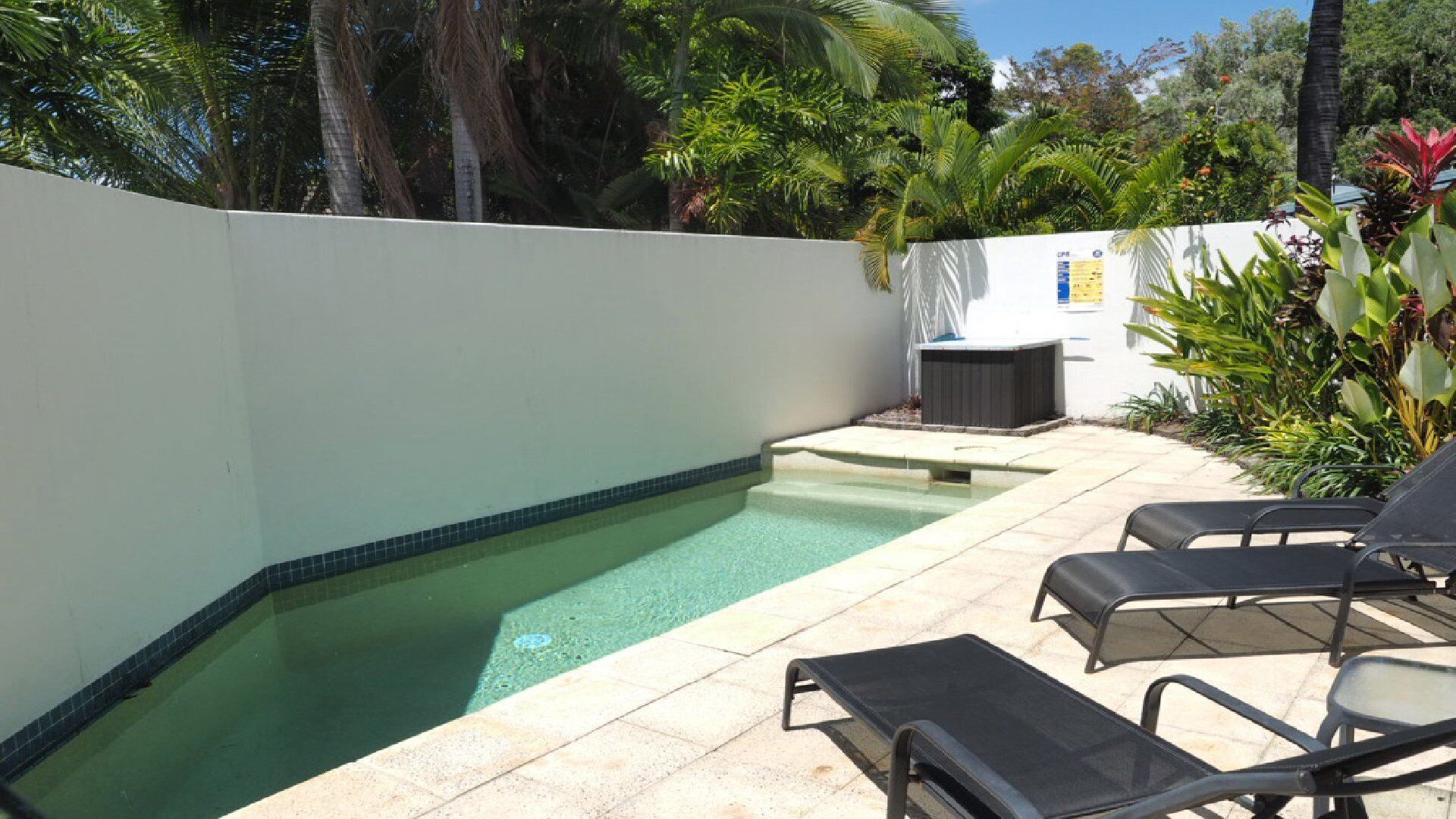Ein Pool mit gefliestem Rand, umgeben von einer weißen Mauer und einem Garten mit verschiedenen Pflanzen. Liegestühle und ein Whirlpool sind zu sehen.
