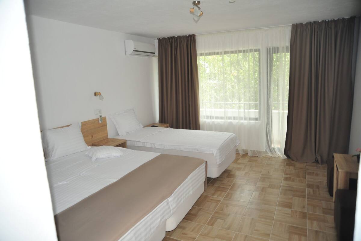 A hotel room with two single beds, a wooden headboard, a bedside table, a nightstand, and a window with curtains.