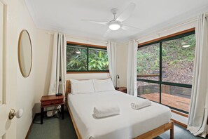 Une chambre à coucher comprenant un lit, une table de chevet, une lampe, un ventilateur de plafond et de grandes fenêtres avec des rideaux.