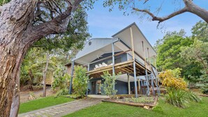 Une maison moderne à deux étages, dotée d’un espace extérieur couvert, et entourée d’une végétation luxuriante et d’arbres.