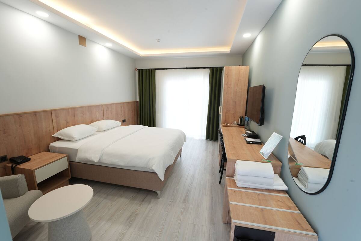 A modern hotel room with a bed, a desk, a chair, a small table, a mirror, and a TV.