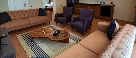 Una sala moderna con un sofá, sillones y una mesa de centro. Se observa una zona de cocina con armarios de madera y una encimera.