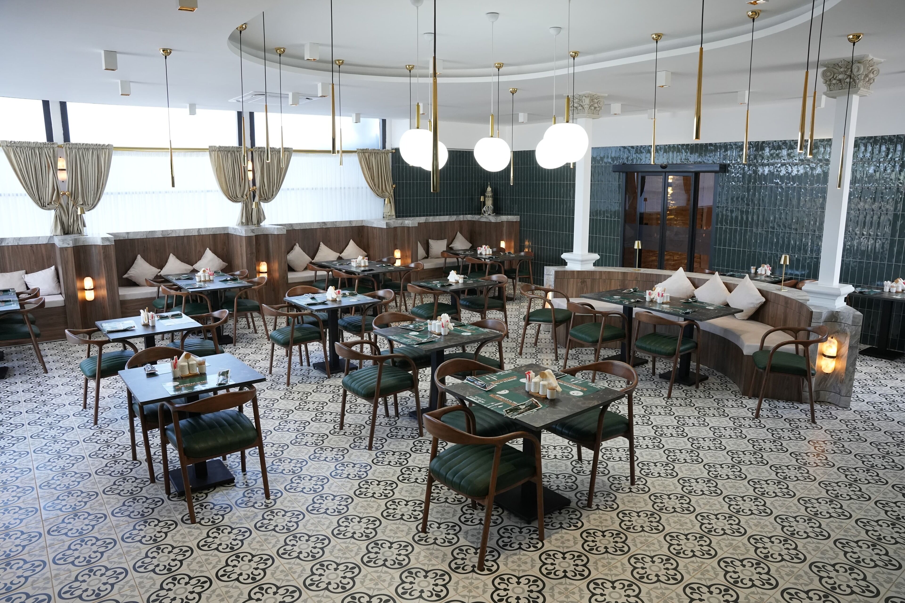 A restaurant with round tables, green cushioned chairs, and patterned floor tiles.
