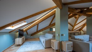 Une chambre aménagée dans les combles, avec un plafond en pente, des poutres apparentes, une télévision, un canapé, une petite table et un tapis à motifs.