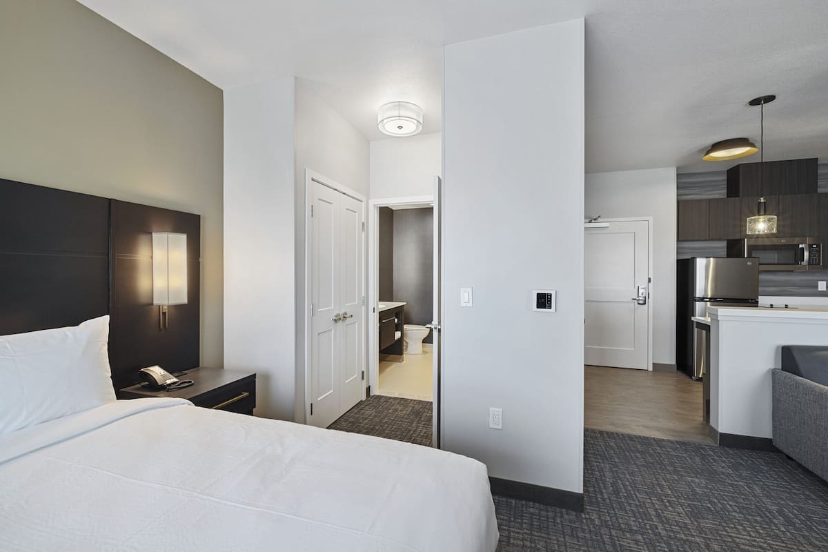 A hotel room with a bed, bedside table, and a view into the kitchen area.
