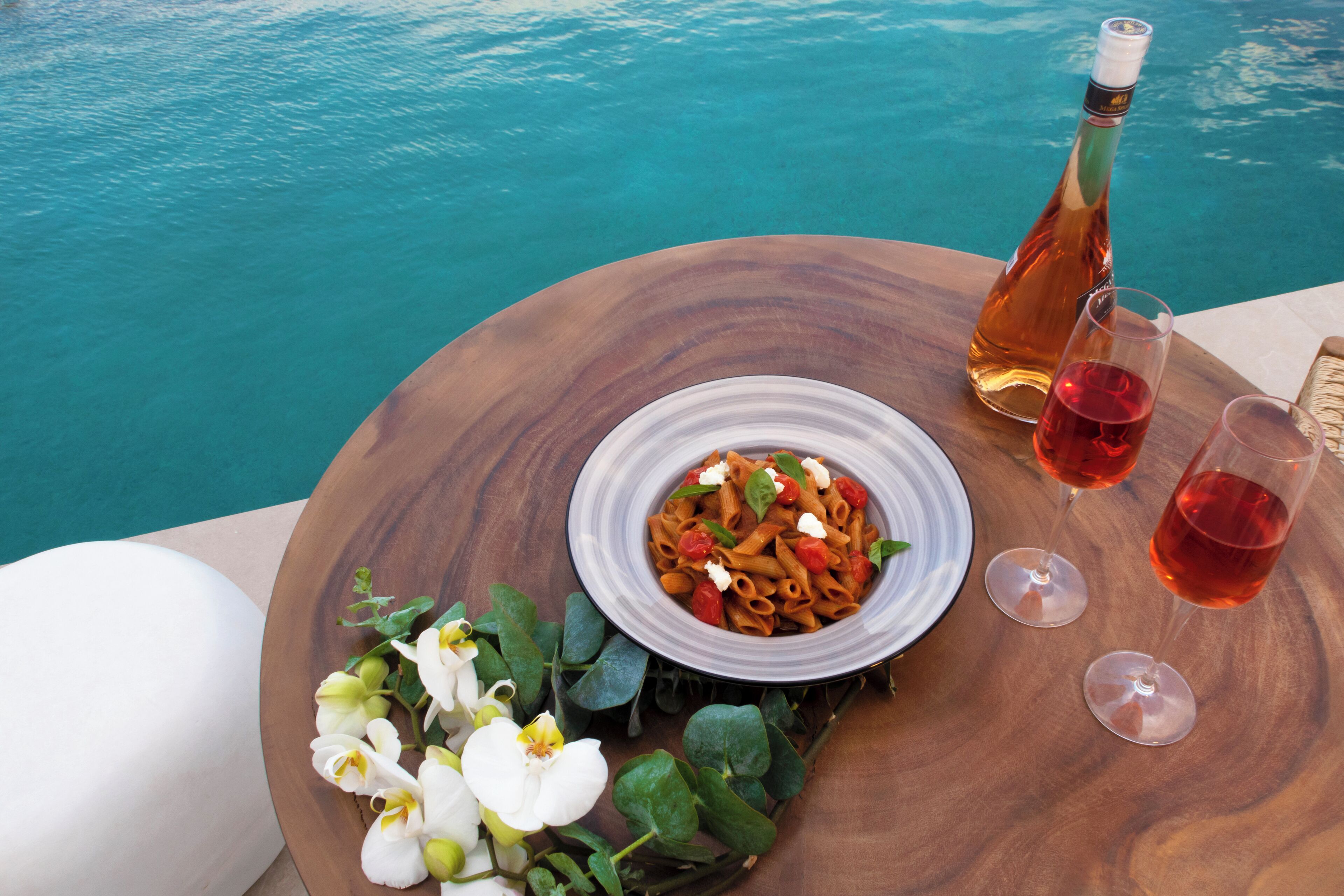 Una mesa de madera con un plato de pasta, dos copas de vino tinto y una botella de vino junto a una piscina.