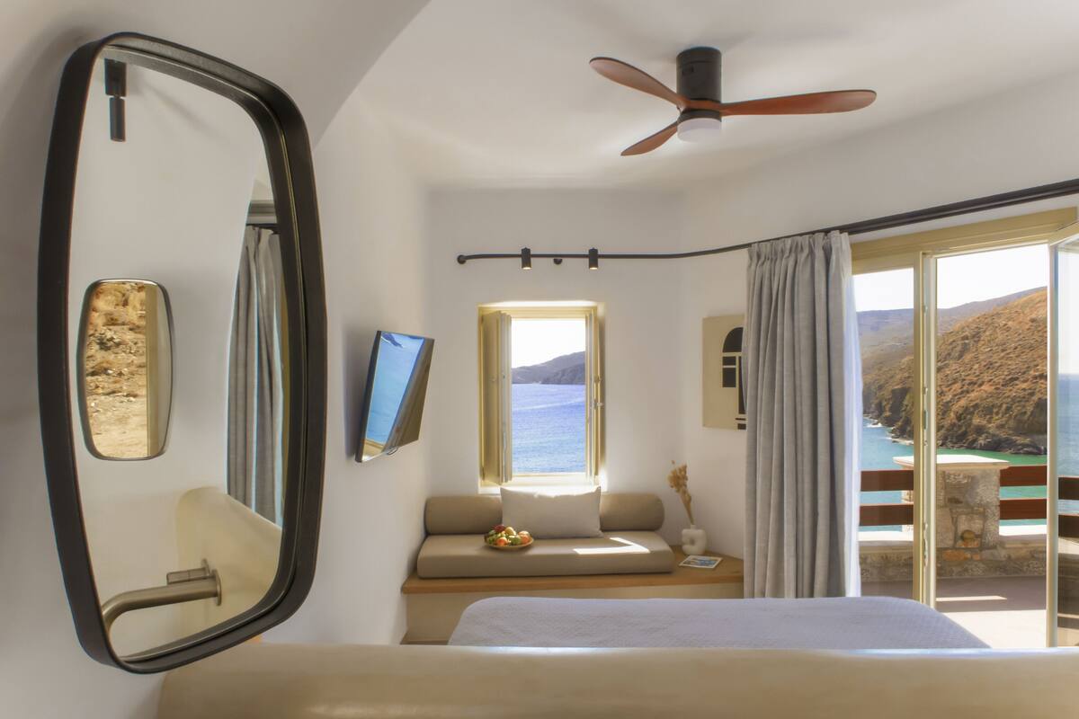 Un dormitorio con cama, un espejo grande, un ventilador de techo, una ventana con vistas al mar y un balcón con mesa y sillas.