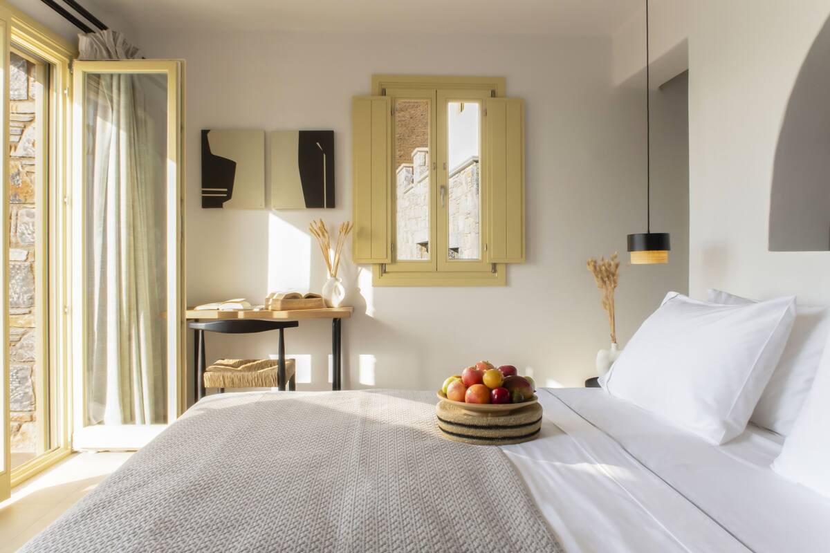 Un dormitorio con cama, una mesita de noche con un frutero, una ventana con cortinas y una lámpara fijada a la pared.