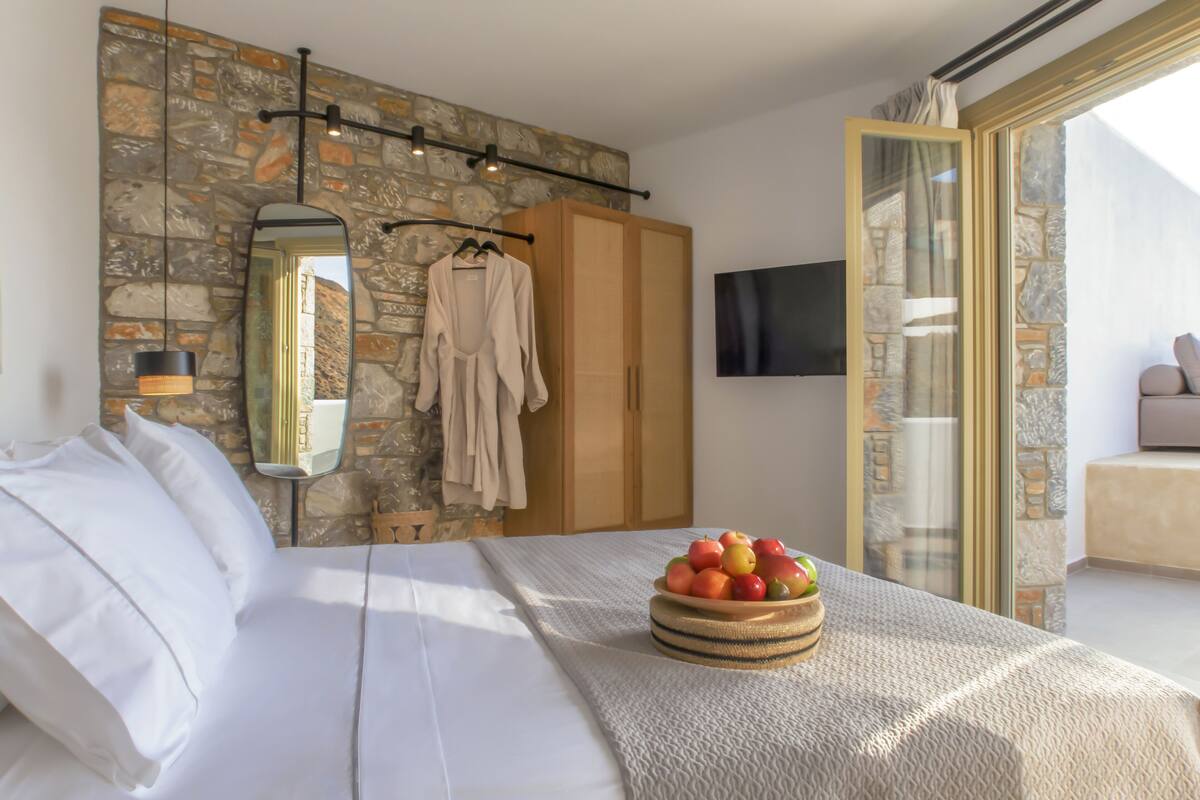 Un dormitorio con pared de piedra, cama con sábanas blancas, frutero sobre la cama, bata colgada y televisor montado en la pared.