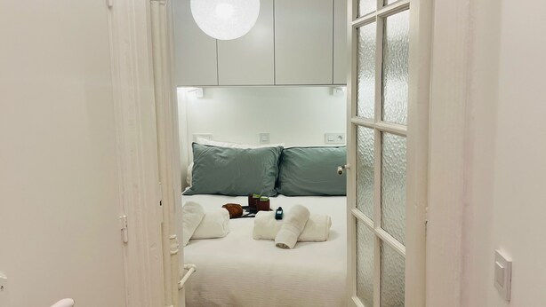 A narrow hallway leading to a bedroom with a bed, pillows, and a hanging light fixture. - Standing - Place des Vosges - Chambre au Calme (Paris)