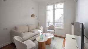A modern living room with a sofa, two armchairs, a coffee table, a TV, and a large window with a view of buildings. - Standing - Place des Vosges - Chambre au Calme (Paris)