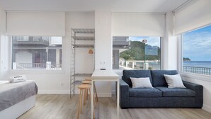 Een moderne, minimalistisch ingerichte kamer met een bed, een grijze bank met kussens, een klein houten tafeltje en uitzicht op een kustlandschap door grote ramen.