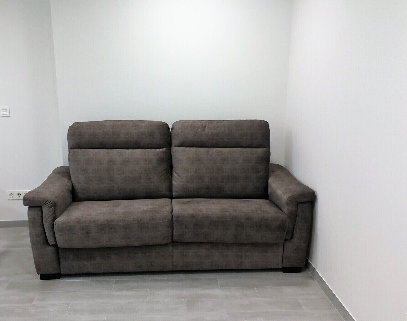 A grey sofa in a corner of a room with white walls and grey flooring.