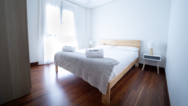 Een slaapkamer met een houten bed, witte beddengoed, een nachtkastje met lamp en een raam met dunne gordijnen.