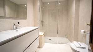 Een moderne badkamer met een douche in een glazen cabine, een witte wastafel met zeepdispenser, en een toilet met een handdoek erop.