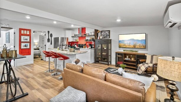 A modern living room with a brown leather sofa, a dining area with red bar stools, a kitchen with red accents, and a mounted TV displaying a landscape. - Renovated Bungalow with Work Space, Kitchen, Patio (St. Augustine)