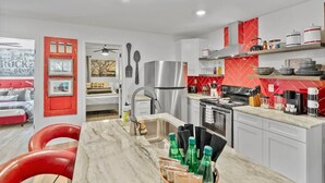 A modern kitchen with a stainless steel refrigerator, a sink, and a countertop with kitchen utensils. There is a dining area with a red chair and a table. - Renovated Bungalow with Work Space, Kitchen, Patio (St. Augustine)