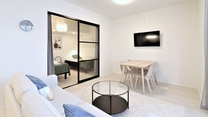 Uma sala moderna com um sofá, uma mesa de centro de vidro, uma mesa de jantar com cadeiras, uma TV fixada na parede e uma porta de vidro deslizante que dá para um quarto.