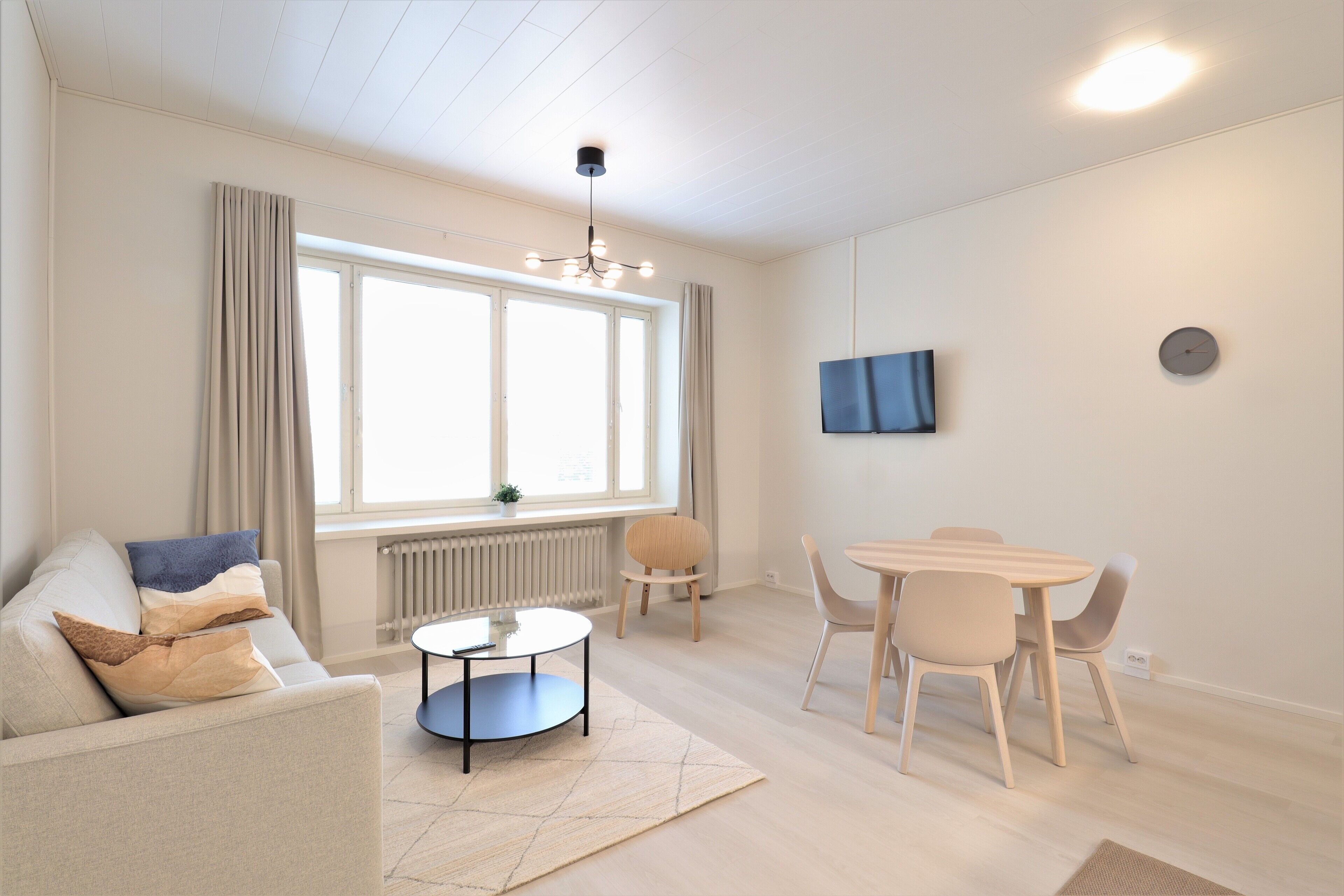 A modern living room with a sofa, coffee table, dining table, and chairs. There is a large window with curtains, a radiator, and a mounted TV on the wall.