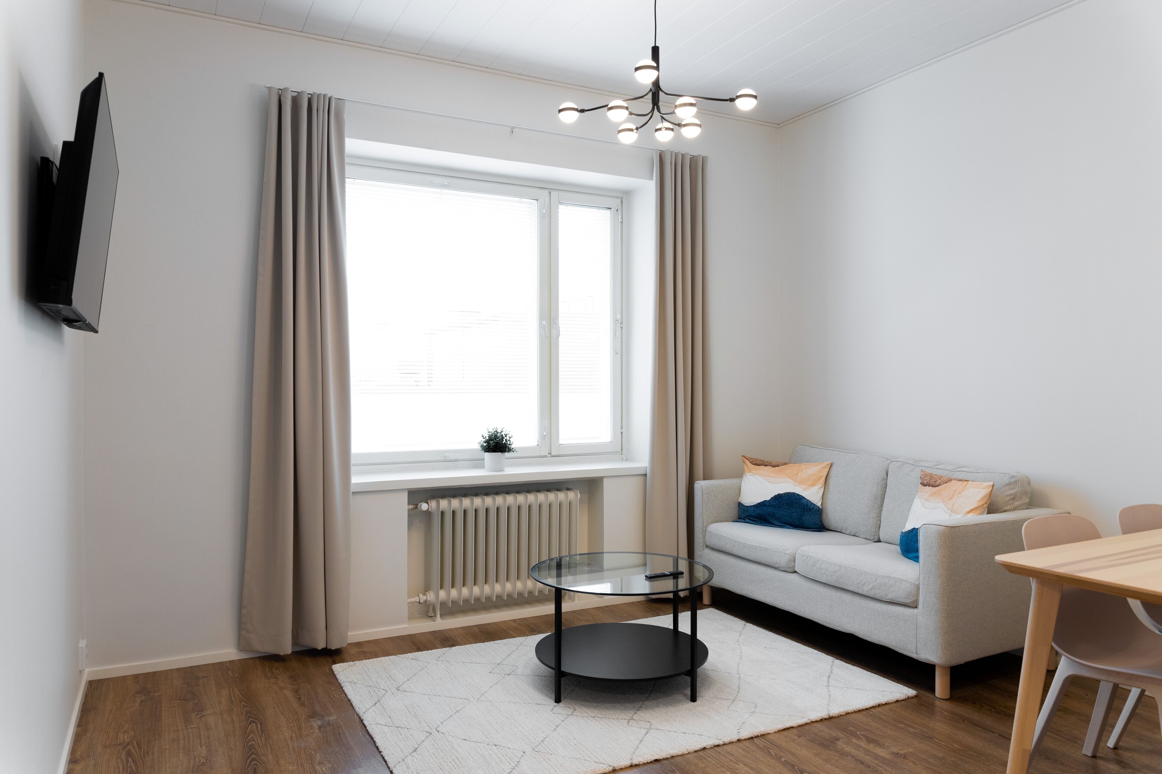 A modern living room with a sofa, a glass coffee table, a TV mounted on the wall, and a window with curtains.