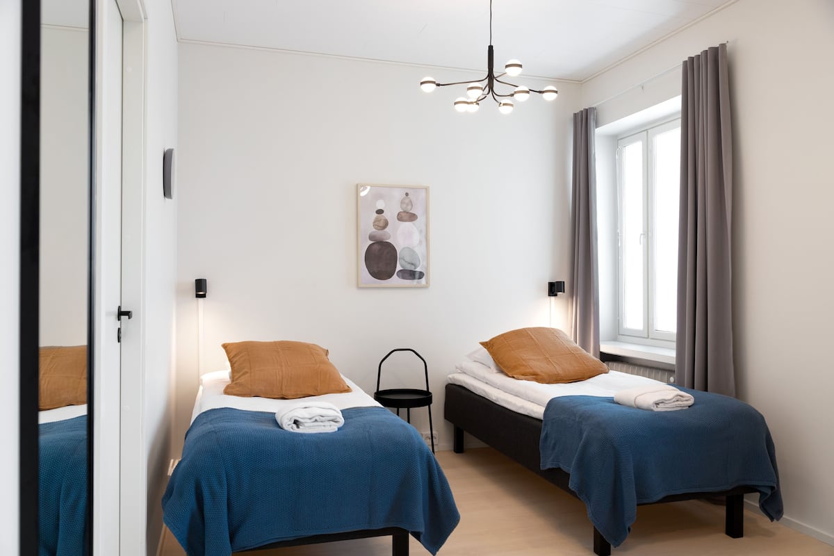 A modern bedroom with two beds, a small side table, a wall-mounted light fixture, and a window with curtains.