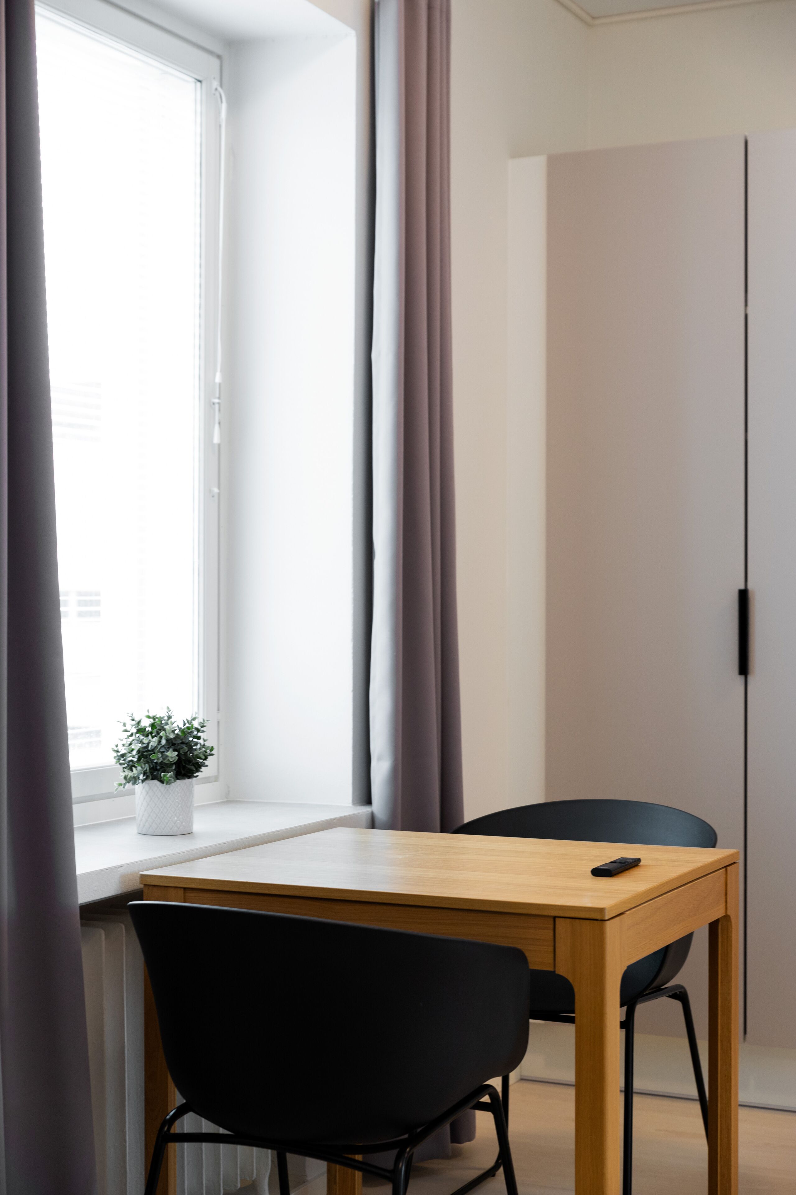 A room with a wooden table and two black chairs, a window with curtains, and a potted plant on the windowsill.