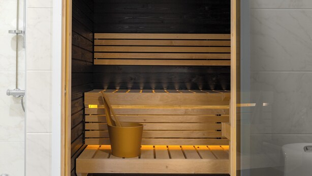 Uma sauna com bancos de madeira e um assento de banco, um balde de metal e uma porta de vidro.