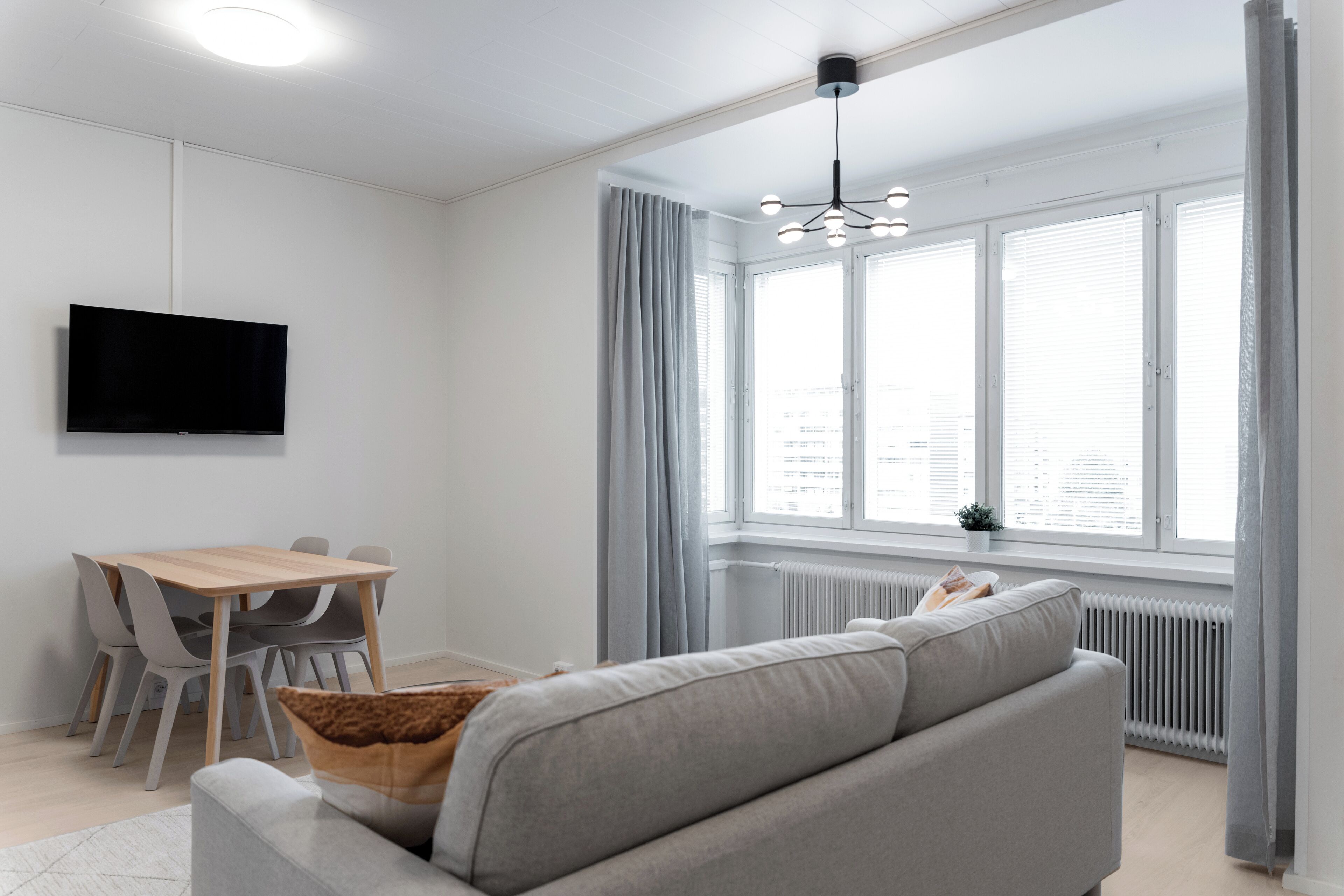 A modern living room with a grey sofa, a wooden dining table with chairs, a flat-screen TV mounted on the wall, and a large window with curtains.