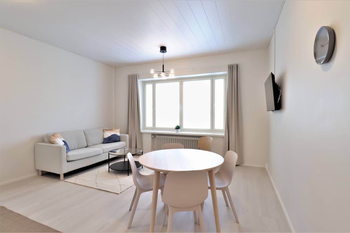 A modern living room with a sofa, a round dining table with chairs, a hanging light fixture, a large window with curtains, and a wall-mounted TV.