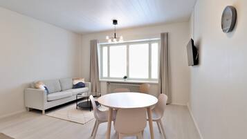 A modern living room with a sofa, a round dining table with chairs, a hanging light fixture, a large window with curtains, and a wall-mounted TV.