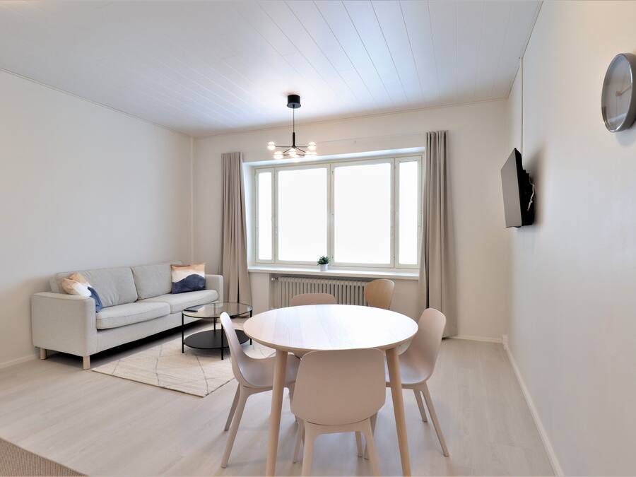 Un salon moderne comprenant un canapé, une table de salle à manger ronde avec des chaises, un luminaire suspendu, une grande fenêtre avec des rideaux et un téléviseur fixé au mur.