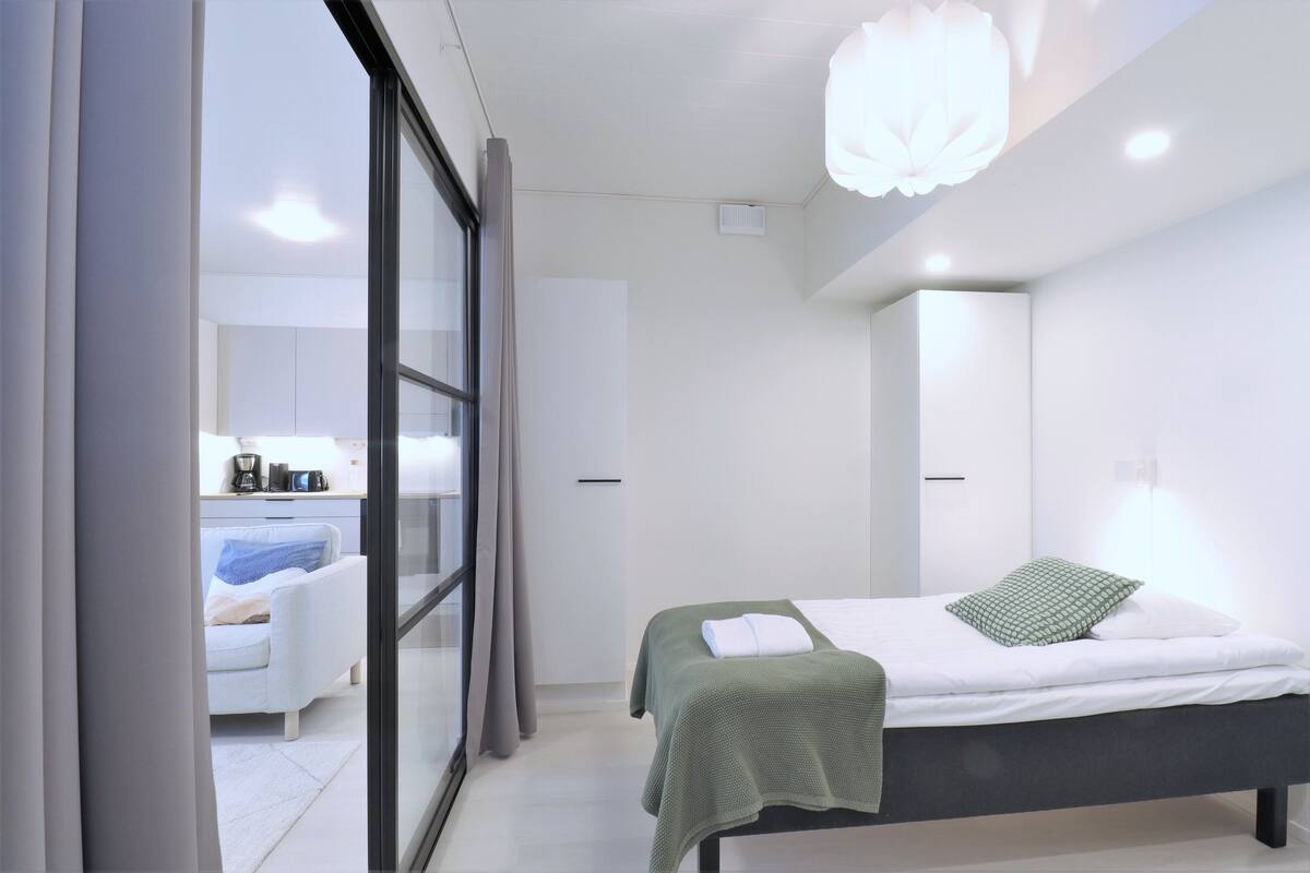 A modern bedroom with a bed, a bedside table, a large window, and a view of a bathroom.