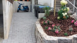 A paved pathway leading to a residential entrance with a scooter parked nearby, a small garden with flowers, and a stone retaining wall. - Moderne Ferienwohnung mit Eigener Terrasse (Arnsberg)