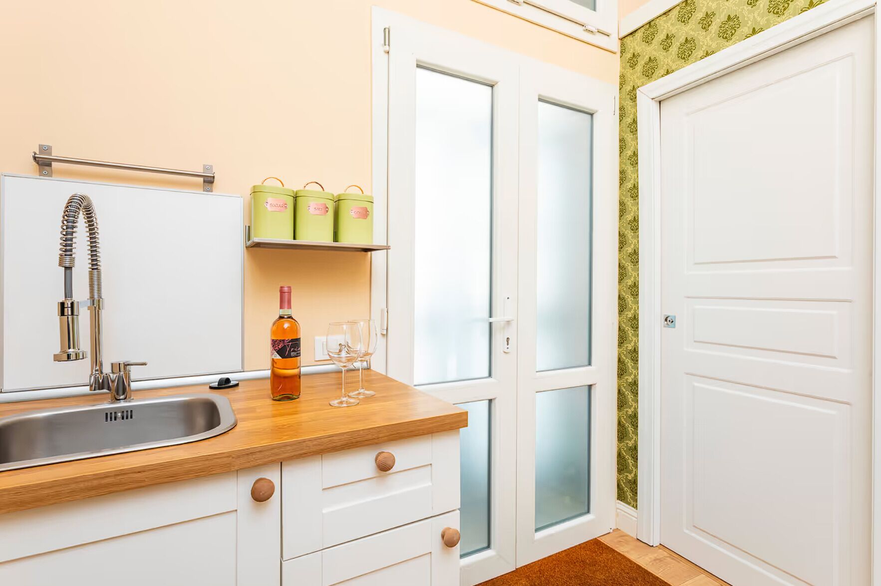 A kitchen with a stainless steel sink, a wine bottle, and two wine glasses on a wooden countertop. There are three green storage containers on a shelf above the counter.