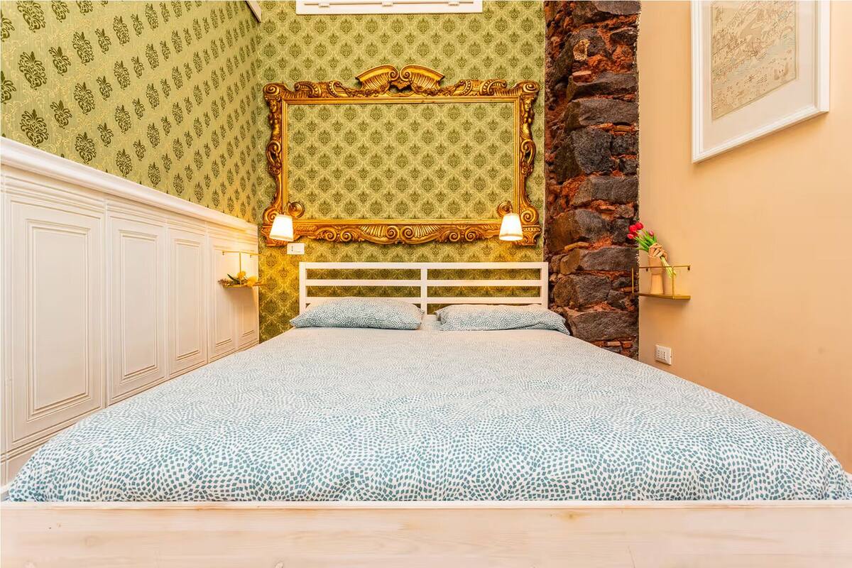 A bedroom with a patterned wallpaper, a decorative headboard, two bedside lamps, and a stone accent wall.